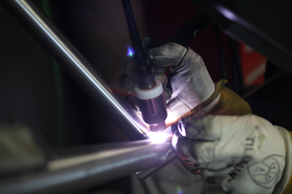 Here’s a close-up of TIG welding the down tube to the bottom bracket shell. For the geeks, TIG stands for Tungsten Inert Gas. The brightly glowing electrode is the tungsten, which provides the arc. The inert gas is argon, which after emerging from the pinkish colored gas lens forms a protective cloud (invisible) around the […]